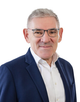 Mann mit grauen Haaren und Brille steht frontal und blickt in die Kamera; trägt dunkelblaues Sakko und weißes Hemd vor neutralem, weißem Hintergrund, Halbporträt ohne sichtbare Zusatzobjekte.