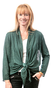 Erwachsene Person mit blondem Haar steht, blickt in die Kamera; eine Hand in der Tasche, andere an der Hüfte; grünes, vorn geknotetes Jäckchen über weißem Top, schwarze Hose, weißer Hintergrund.