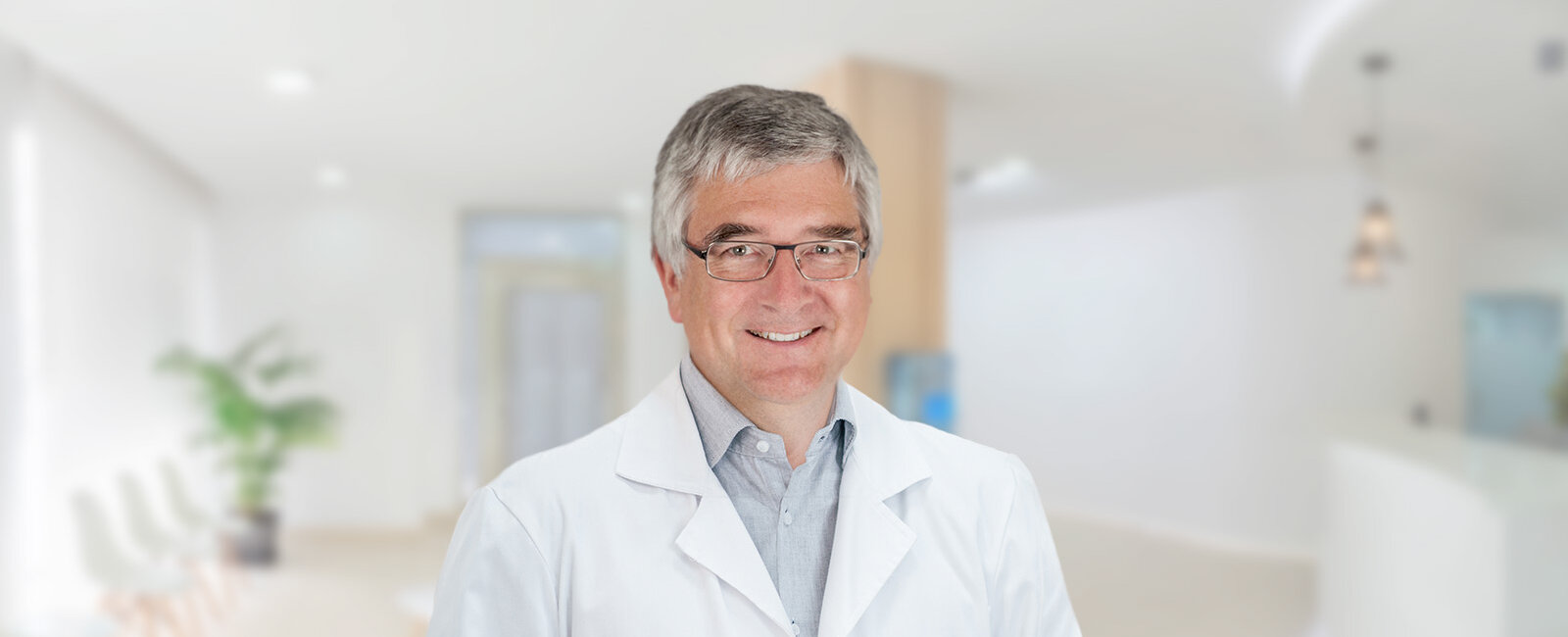 Prof. Dr. Joachim Grfika mit grauen Haaren, Brille und weißem Kittel lächelt in die Kamera; im Hintergrund ein heller, unscharfer Innenraum mit Wartebereichsstühlen, Pflanze, Empfangstresen, Pendelleuchten und Glastür.