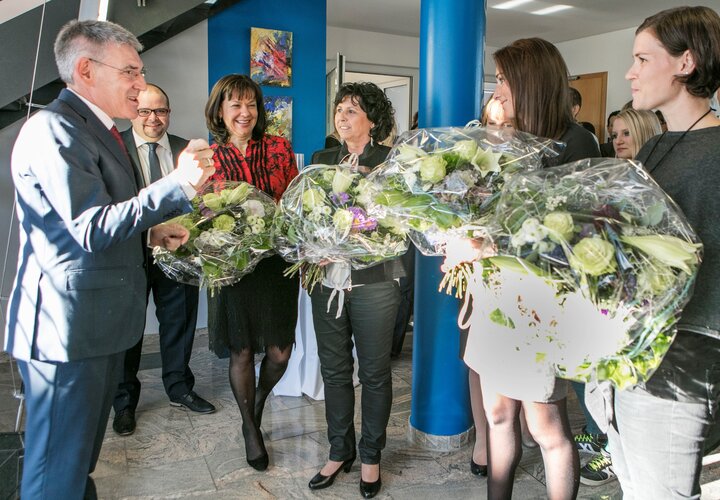 Gruppe Erwachsener in Businesskleidung; ein Mann links spricht und gestikuliert, mehrere Frauen halten große, in Folie verpackte Blumensträuße; helle Innenlobby mit blauer Säule, abstrakten Gemälden und weiteren Personen.