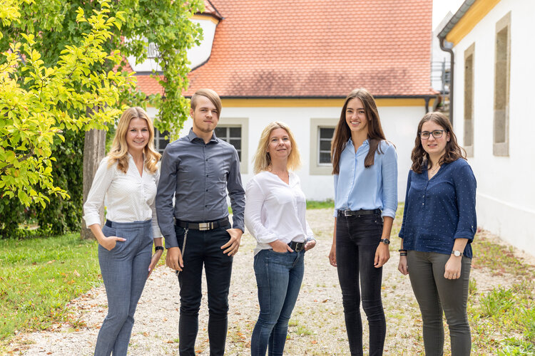 Fünf Personen stehen nebeneinander und blicken in die Kamera, auf einem Kiesweg in einem Hof zwischen weißen Gebäuden mit roten Ziegeldächern; links grüner Baum, helles Tageslicht.