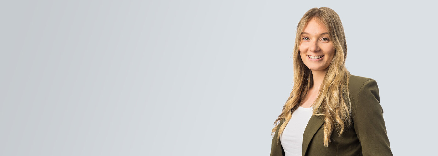 Erwachsene Person mit langen blonden Haaren im olivgrünen Blazer und weißem Oberteil lächelt in Halbporträt zur Kamera; rechts ausgerichtet vor hellem, grauem Verlaufshintergrund mit viel Leerraum links.