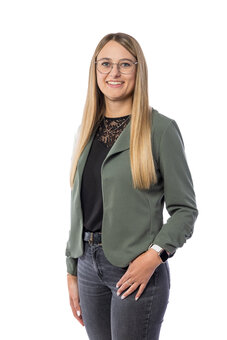 Stehende Person mit langen blonden Haaren und Brille lächelt in die Kamera, trägt grünen Blazer, schwarzes Oberteil mit Spitzenbesatz, graue Jeans und Smartwatch; Studioaufnahme vor weißem Hintergrund.